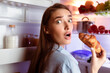 © Prostock-studio - Cheat meal concept. Scared young woman eating croissant near fridge late in night