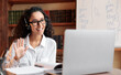 © Prostock-studio - Distance Education. Positive lady wearing glasses and wireless headest at virtual meeting, sitting at desk, having video call on laptop, waving to webcam. Woman studying or teaching online at home
