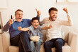 © Prostock-studio - Soccer Fans. Happy Son, Father And Grandpa Watching Sport Match On TV Celebrating Victory Sitting On Sofa At Home.