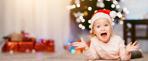 Wall Mural - Christmas surprise. Happy emotional little girl in Santa hat lying on floor at home, opened mouth in excitement