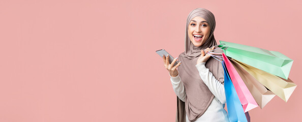 Wall Mural - Online shopping and cashback concept. Cheerful muslim girl in hijab holding smartphone and bright paper bags, enjoying seasonal sales, pink background