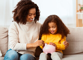 family savings. cheerful afro mother teaching little daughter how to save money holding piggybank si