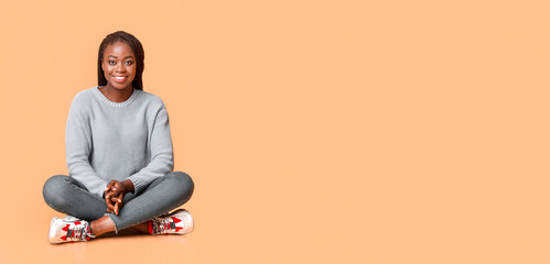 Peaceful smiling black woman sitting on floor, beige background, copy space, panorama