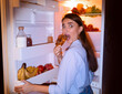 © Prostock-studio - Midnight snack. Scared girl in pajamas eating croissant near opened fridge, free space