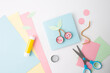 © Elena - Flat lay of craft materials on white surface: pink, blue, green paper, blue-handled scissors, yellow glue stick, pink cherry cut outs with faces, green leaf on light blue square.