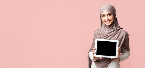 Wall Mural - Muslim girl in headscarf demonstrating digital tablet with black screen over pink background in studio, empty space