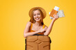 © Prostock-studio - Happy girl tourist lean on suitcase and holding flight tickets and map, ready to travel, posing over yellow background with copy space