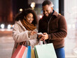 © Prostock-studio - App For Online Shopping. Portrait of smiling black man and woman using mobile phone, lady pointing at screen, couple holding shopper bags, standing outdoors in the evening, choosing gift