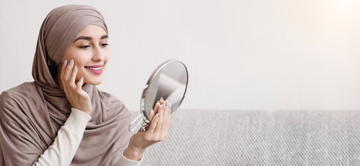 Wall Mural - Beauty routine. Attractive muslim girl in hijab holding mirror and looking at her face with perfect soft skin, sitting on sofa at home, free space