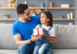 © Prostock-studio - This Present Is For You. Portrait of loving girl sitting on dad's lap and greeting him with father's day or birthday, holding wrapped gift box, happy family celebrating holiday together at home