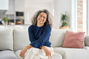 happy middle aged woman in her 50s sitting on sofa in living room at home. smiling beautiful mature 