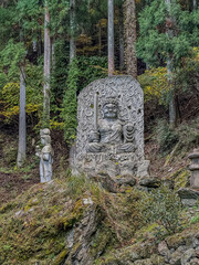 A serene Buddhist stone statue in a forest setting during autumn, surrounded by lush green trees and mossy rocks, creating a peaceful, spiritual atmosphere.