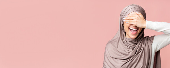 Wall Mural - Happy Muslim Girl In Hijab Covering Eyes With Hand And Smiling, Having Fun Over Pink Background, Free Space