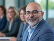 © MindVerse - A business meeting with businessmen and businesswomen around the tables looking to camera, a middle aged man in foreground is focused and people on background are defocused with office area
