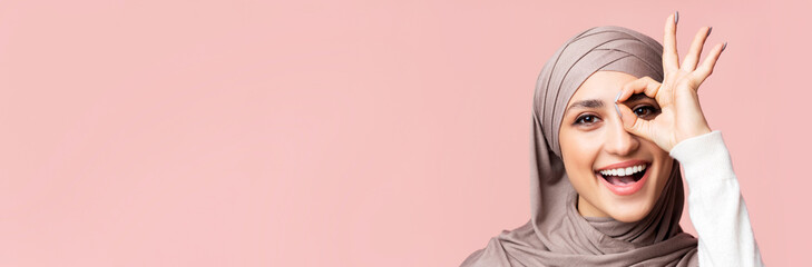 Wall Mural - Cheerful arabic lady in hijab showing ok gesture, holding hand near eye, looking through fingers at camera, pink background with empty space, closeup