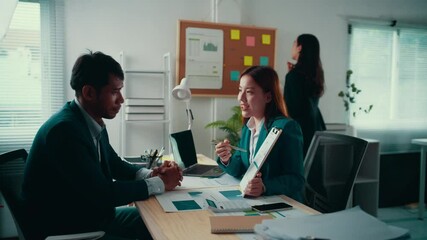 Sticker - Businesswoman is showing and explaining something on a document to her male colleague while sitting at a desk in an office. Another woman is walking in the background