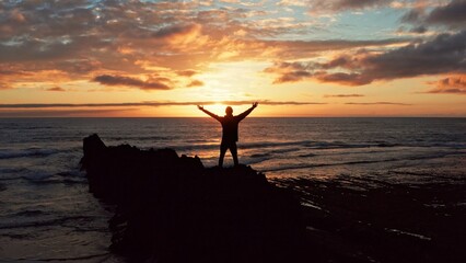 Wall Mural - Man stands on a rock against the ocean during summer sunset. Human with raised hands looks to sun over horizon in morning while sunrise. Man feels happy and freedom. Silhouette of a man against  sky