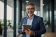 © Tamii - Portrait of happy 50 years old business man executive standing in office using digital tablet. Middle aged businessman manager business owner working on professional finances project looking at camera