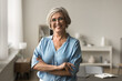 © fizkes - Smiling mature woman posing alone with arms-crossed indoor, standing in homeoffice or cozy living room, exude professionalism and confidence. Individual entrepreneur, home portrait of happy retiree