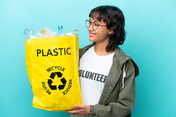 Wall Mural - Young Argentinian woman holding a bag full of plastic bottles to recycle isolated on blue background looking side