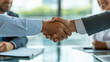 © Anucha - Close-up of handshake between two business professionals in a conference room, indicating a successful business deal or collaboration. Other team members are seated at the back, emphasizing trust.