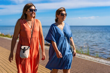 Summer vacation. Portrait of two pretty mid-adult women wearing flowing dresses walking on sidewalk by seaside resort on beautiful sunny day.