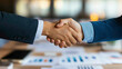 © Anucha - Two business partners shake hands over a table covered with financial reports, sealing a collaborative business agreement. The handshake symbolizes trust, mutual success, and professionalism.