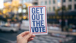 © T. Supisararaviwong - A hand holding flyer that says Get Out Vote in city setting.