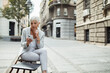 © Davor - Senior businesswoman sitting on bench using smartphone in city