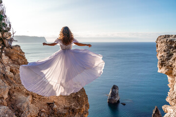 A woman in a white dress stands on a rocky cliff overlooking the ocean. The scene is serene and peaceful, with the woman's dress billowing in the wind. The ocean is calm and blue.