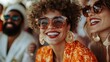 © Dulemegapixel - A woman, beaming with joy, is in vivid orange floral attire and large circular earrings while enjoying time with friends on a sunny day, all sharing smiles together.