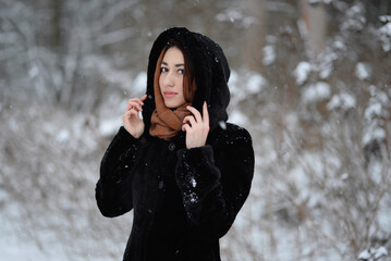 A girl wearing a black coat with a hood posing in a snowy forest in winter. Cold frosty snowy winter weather concept