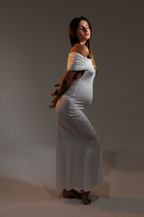 
Beautiful young pregnant brunette girl. A woman waiting for a baby in a white dress with bare shoulders in the studio. Artistic smudges in the photo