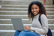 © AI_images_for_people - A cheerful young Black female college student e-learning on her laptop at campus, sitting on the steps and smiling widely at the camera.