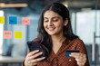 © Liubomir - Businesswoman close-up inside office holding bank credit card and phone, office worker close-up smiling, choosing gifts in online store, and transferring money, booking services remotely.