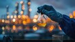 © chutikan - Highlight the gloved hands of a worker holding a bottle with a yellow crude oil sample inside against the backdrop of a refinery.