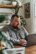 © Vladislava - A plus-size man working on a laptop at a desk, highlighting productivity, technology, and modern work environments, focus and engagement in a home office setting, banner with copyspace for text.