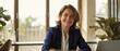© Ai Studio - Professional woman in a suit sits confidently at her desk with a welcoming smile in a sunlit, modern office, exuding warmth and approachability.