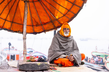 Portrait of an old male indian sadhu baba sitting on ghats near river ganges in a blessing pose at varanasi city in traditional dress under traditional umbrella.
