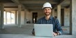 © val - A smiling construction manager in a hard hat works on a laptop at a building site, showcasing professionalism and modern technology in the construction industry.