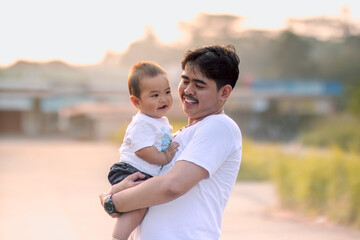 The father held the baby boy warmly and smiled at the baby boy. the baby boy made an adorable smiling expression in the other direction. photo with a natural background with sunlight in the afternoon.