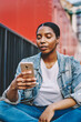 © BullRun - Thoughtful african american teenage female in jeans jacket reading income message on mobile phone sitting outdoors, dark skinned hipster girl connecting her smartphone to 4G internet for blogging
