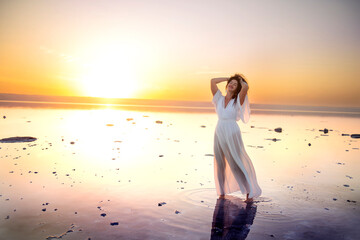 Beautiful girl with long dark curly hair in a long airy white dress stands in the water of a lake at sunset. mirror reflection of the sky in the surface of the lake. Fantastically beautiful view of a 