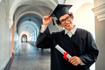 Graduated from college or university. Happy cheerful student in academic gown and mortar board holding diploma with a red ribbon. Copy space.