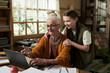 © pressmaster - Elderly woman with white hair in bun is using laptop while young granddaughter embracing her from side in cozy room
