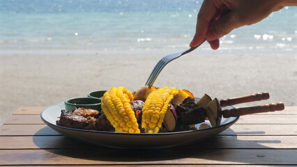 Person eating grilled corn and skewers by seaside, capturing casual meal with ocean view. Summer Vacation and Outdoor Dining.