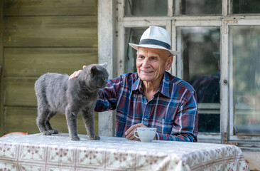 Senior man in a plaid shirt and hat sitting at a table petting a gray cat, smiling outdoors in front of a rustic house