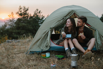 Smiling young man and woman having dinner near a tent in the mountains at sunset.