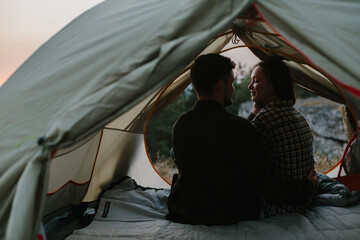 Wall Mural - Smiling young man and woman hugging while sitting in a tent at sunset.