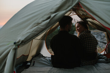 Wall Mural - Smiling young man and woman hugging while sitting in a tent at sunset.